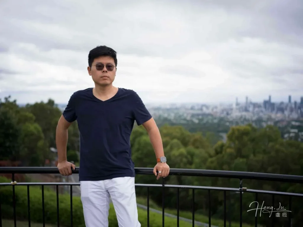 Lu Heng stands at a railing in a dark shirt and white trousers, with trees and a distant city behind him.