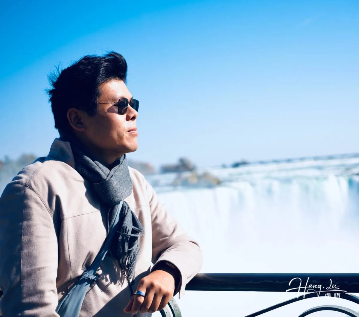 Lu Heng wears sunglasses and a scarf beside a waterfall, with white spray beneath a clear blue sky.