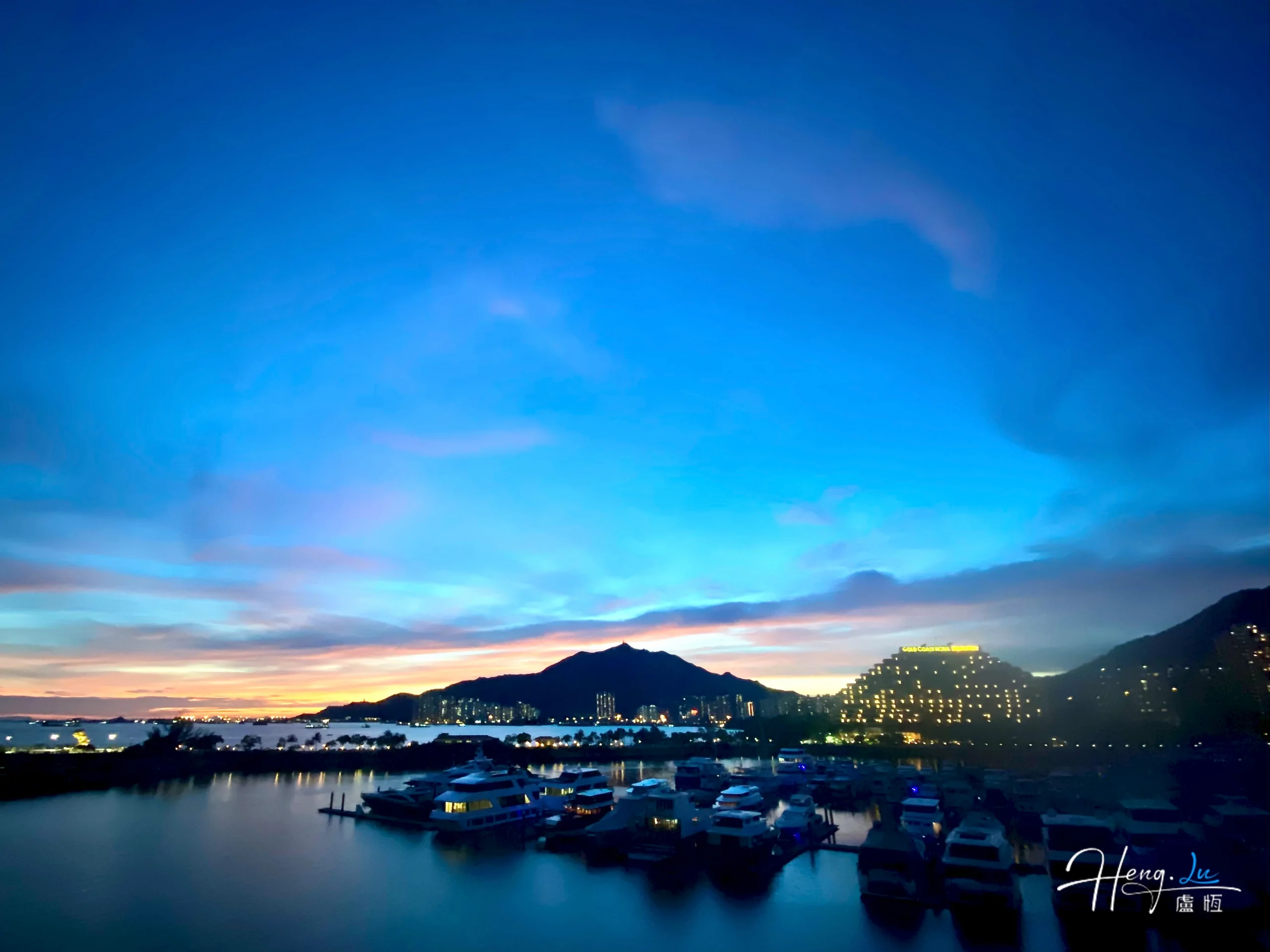 Varios barcos descansan en un puerto bajo un cielo azul de tarde, con luces a lo largo de la orilla.