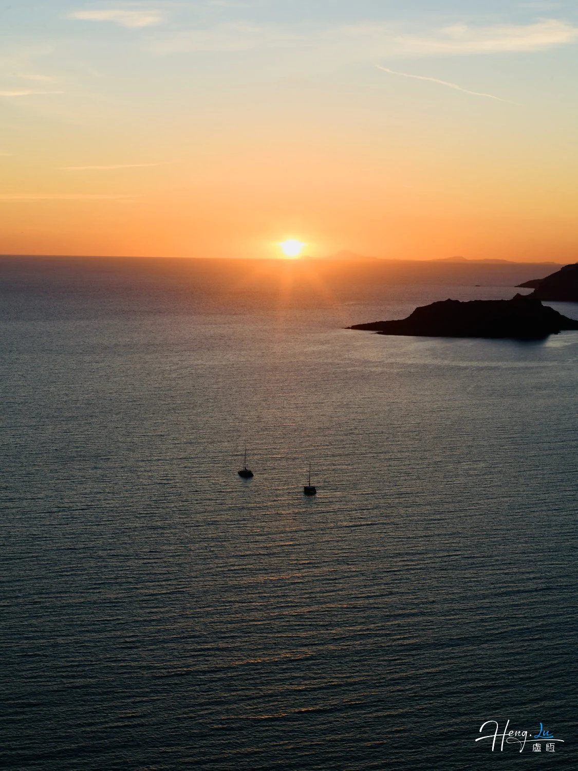 The sun sits above a calm sea, with small boats below and a dark headland to the right.