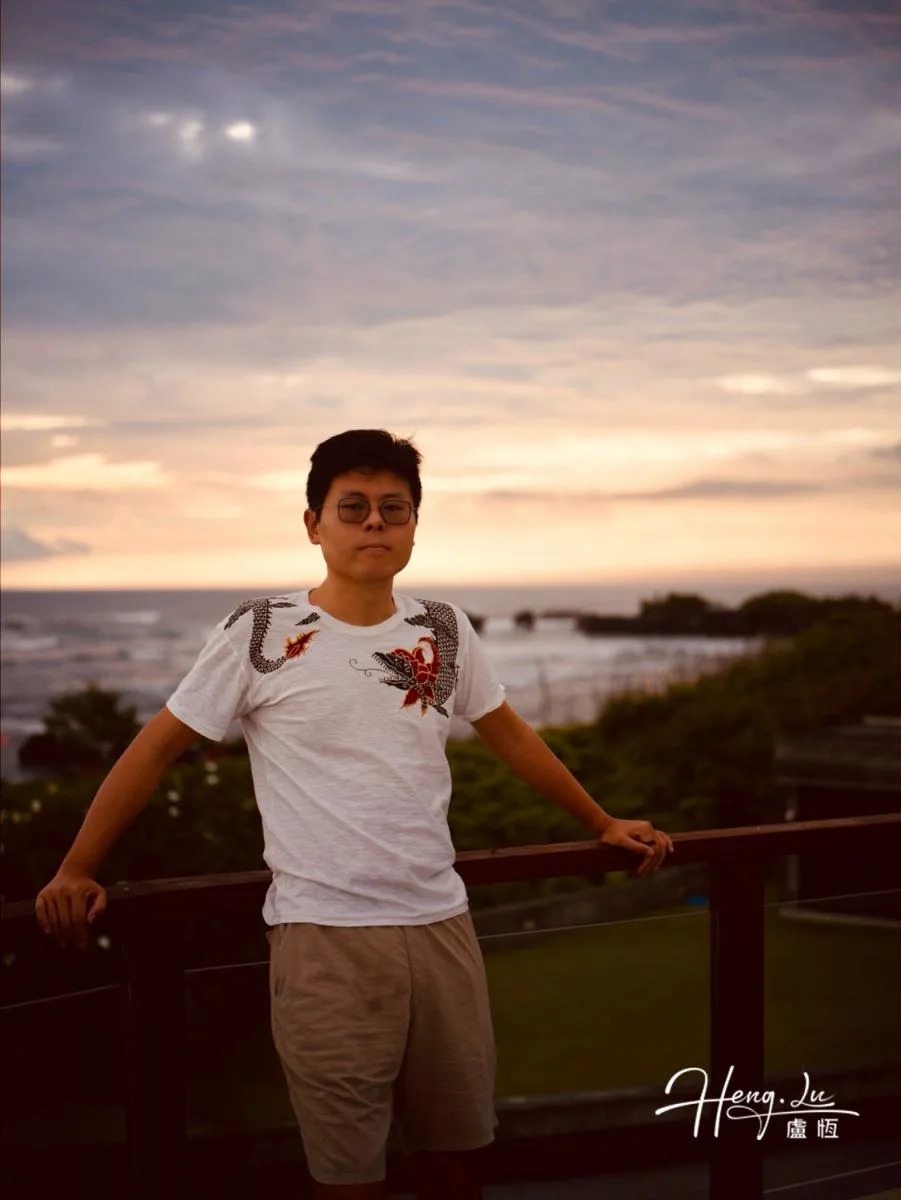Lu Heng rests his hands on a wooden railing, with the sea and a softly lit evening sky behind him.
