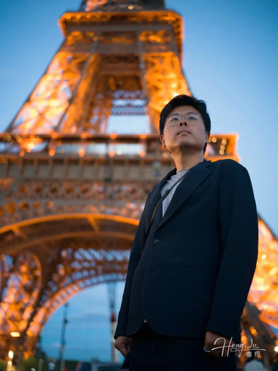 Lu Heng wears a dark jacket beside the illuminated Eiffel Tower as evening turns blue.