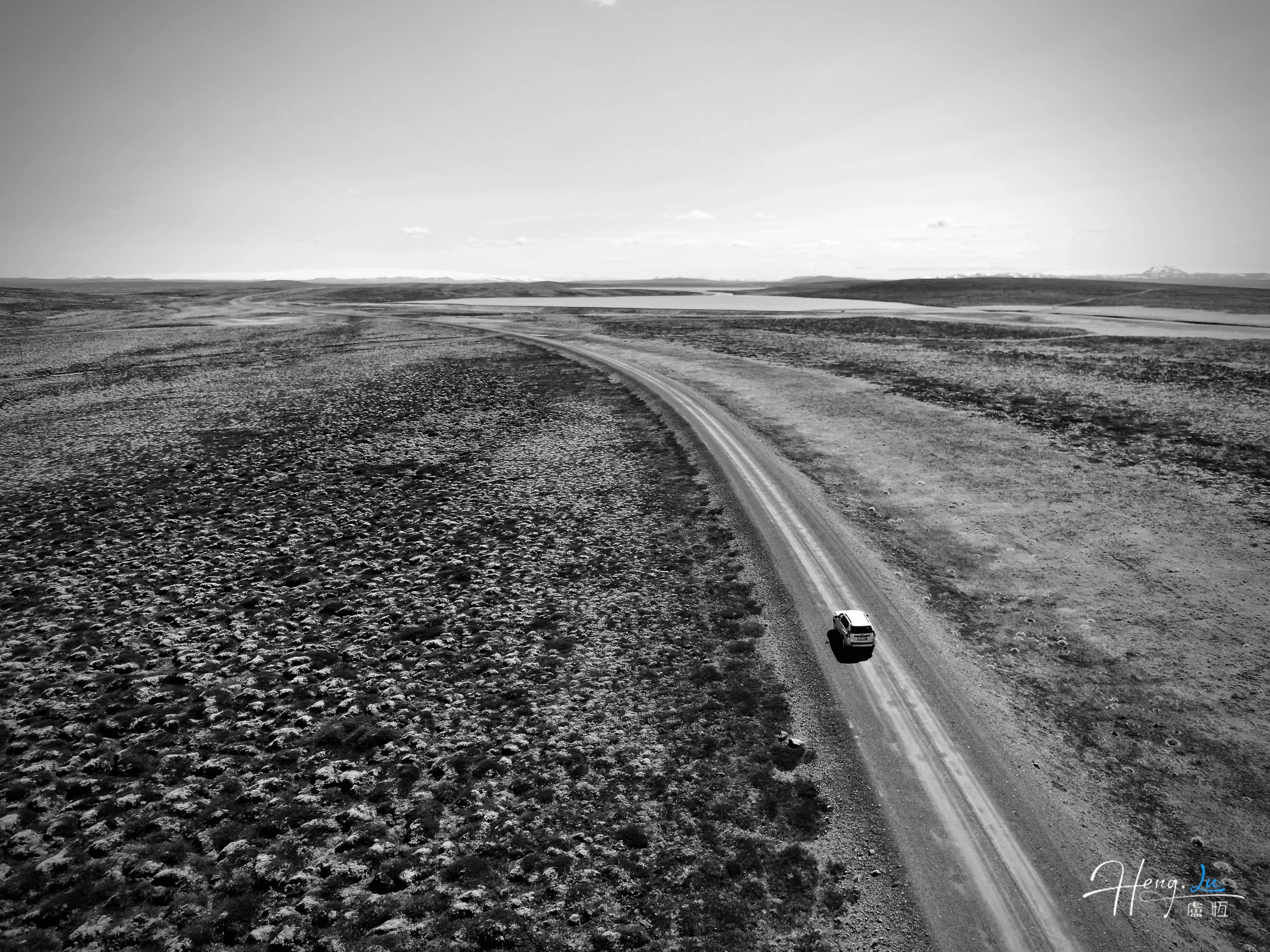 A small car follows a pale road across open, treeless land in a black-and-white aerial photograph.
