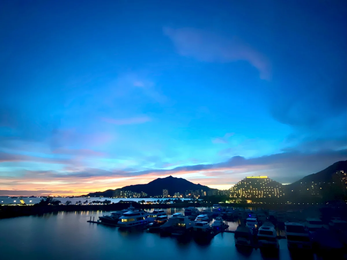 Varios barcos descansan en un puerto bajo un cielo azul de tarde, con luces a lo largo de la orilla.