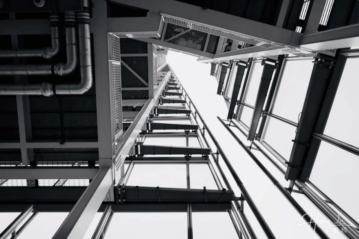 Black-and-white beams, pipes and geometric lines converge towards a bright opening overhead.