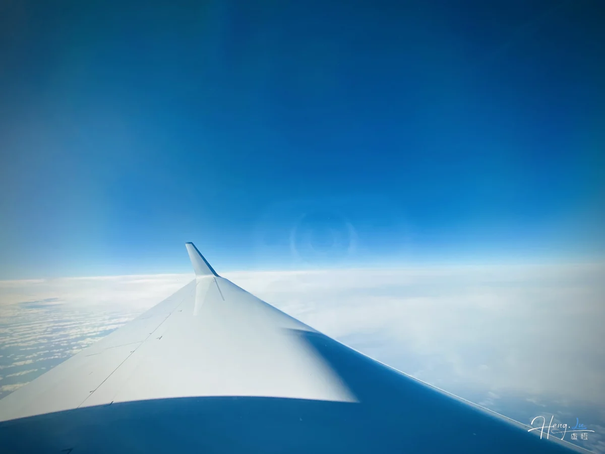 El ala de un avión cruza la parte inferior del encuadre, con una capa de nubes y un amplio cielo azul.