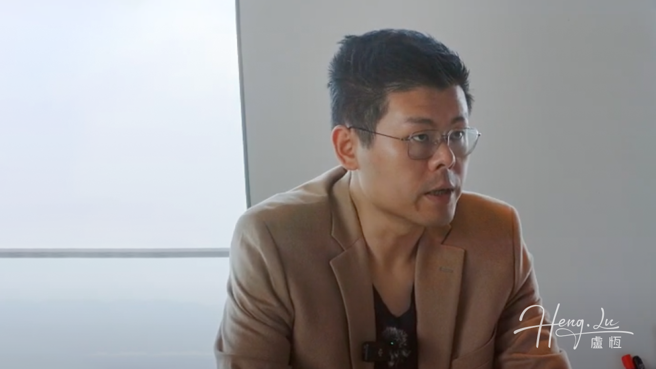 Lu Heng wearing a brown jacket and speaking indoors.