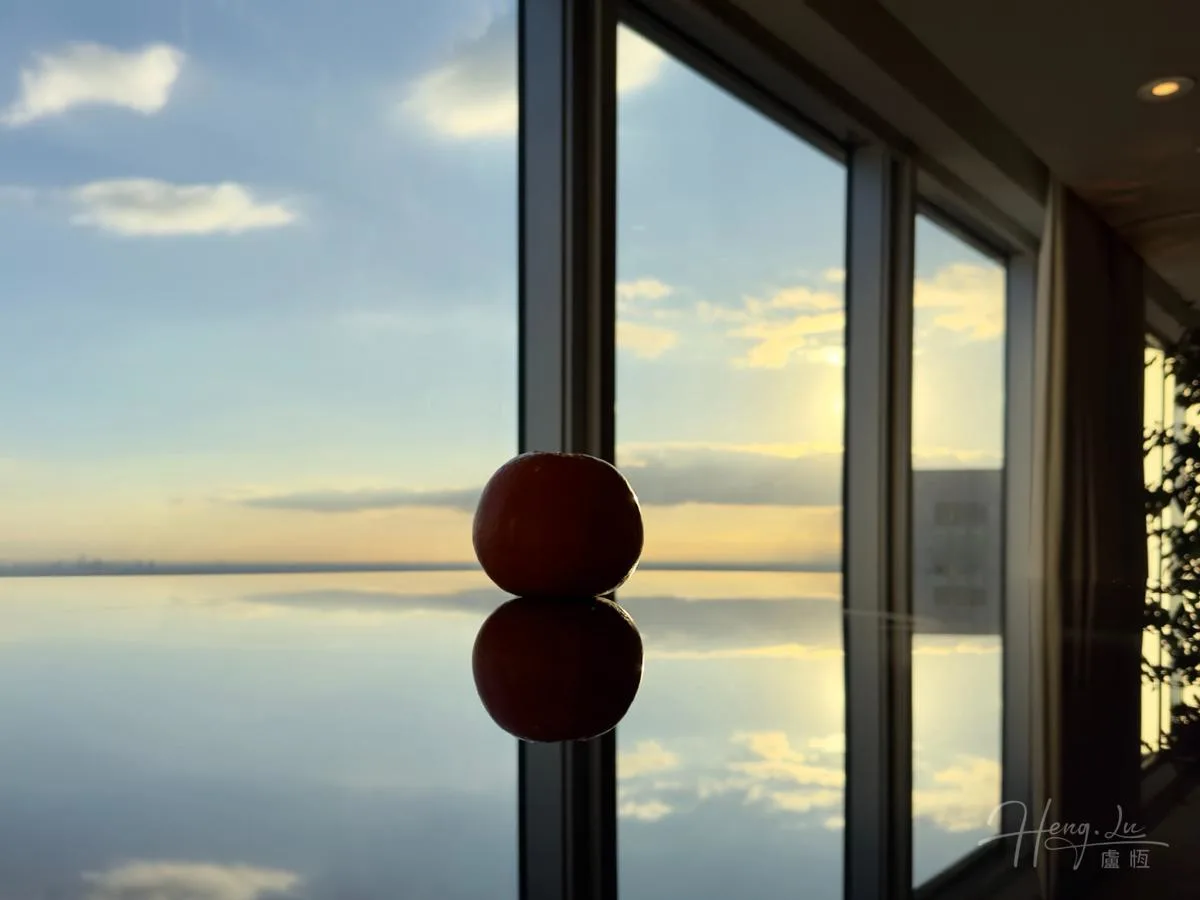 An orange and its reflection sit beside a window, with sky and a distant horizon beyond.