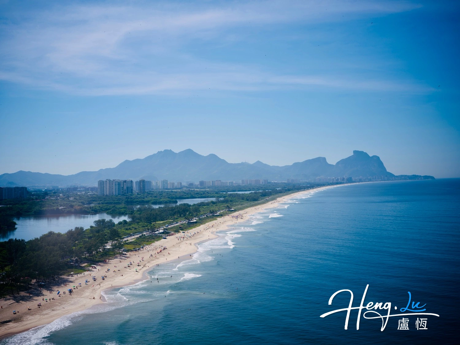 A long beach curves between blue water and a city, with mountains along the horizon.