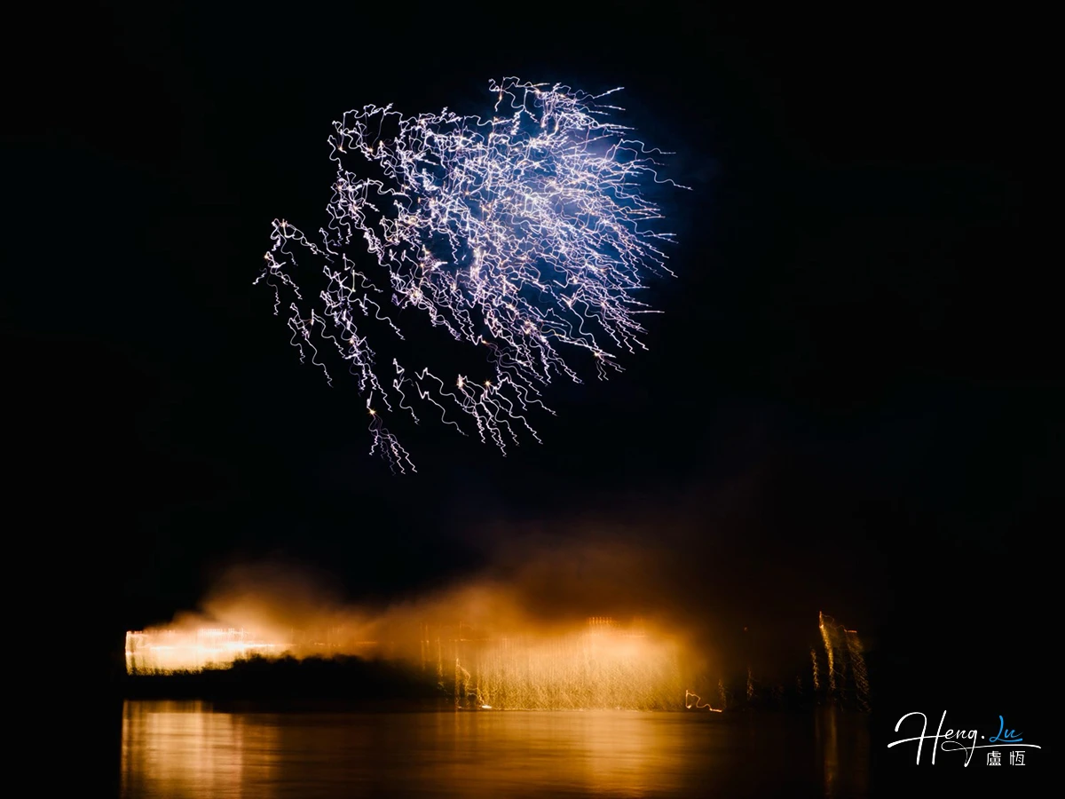 Blue-white firework trails hang above golden light and its reflection on dark water.